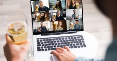 Person raising a glass of beer to a laptop screen of colleagues toasting on a video call
