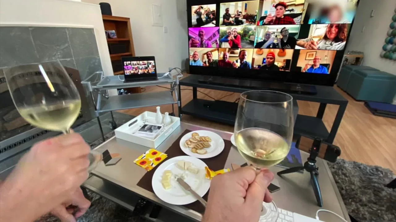 Two glasses of white wine raised in a living room as a TV shows colleagues on a video call