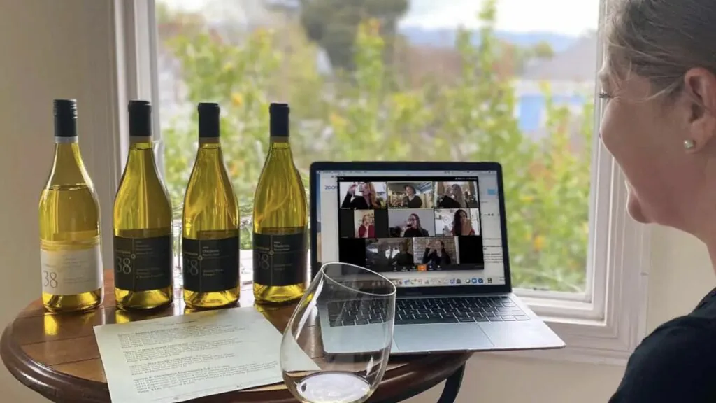 Four white wine bottles and a glass beside a laptop showing a video call grid at a virtual wine tasting