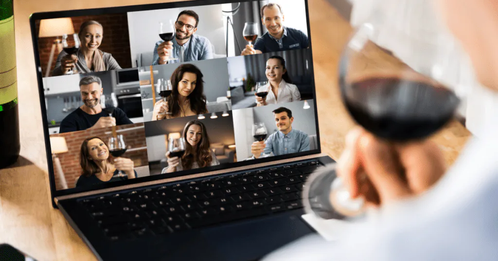 Person holding a glass of red wine while a laptop shows colleagues toasting on a video call