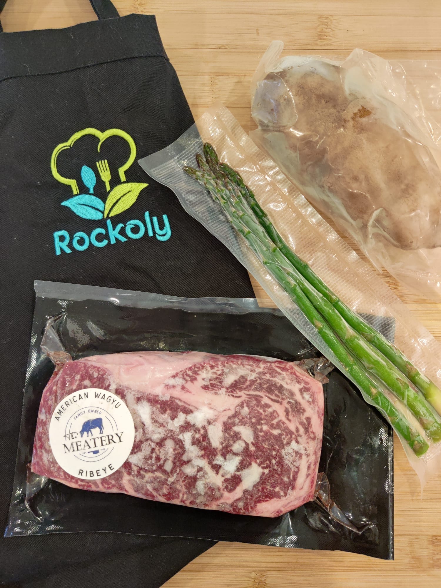 Cooking kit of vacuum-sealed wagyu ribeye, asparagus and potato laid out on a branded Rockoly apron