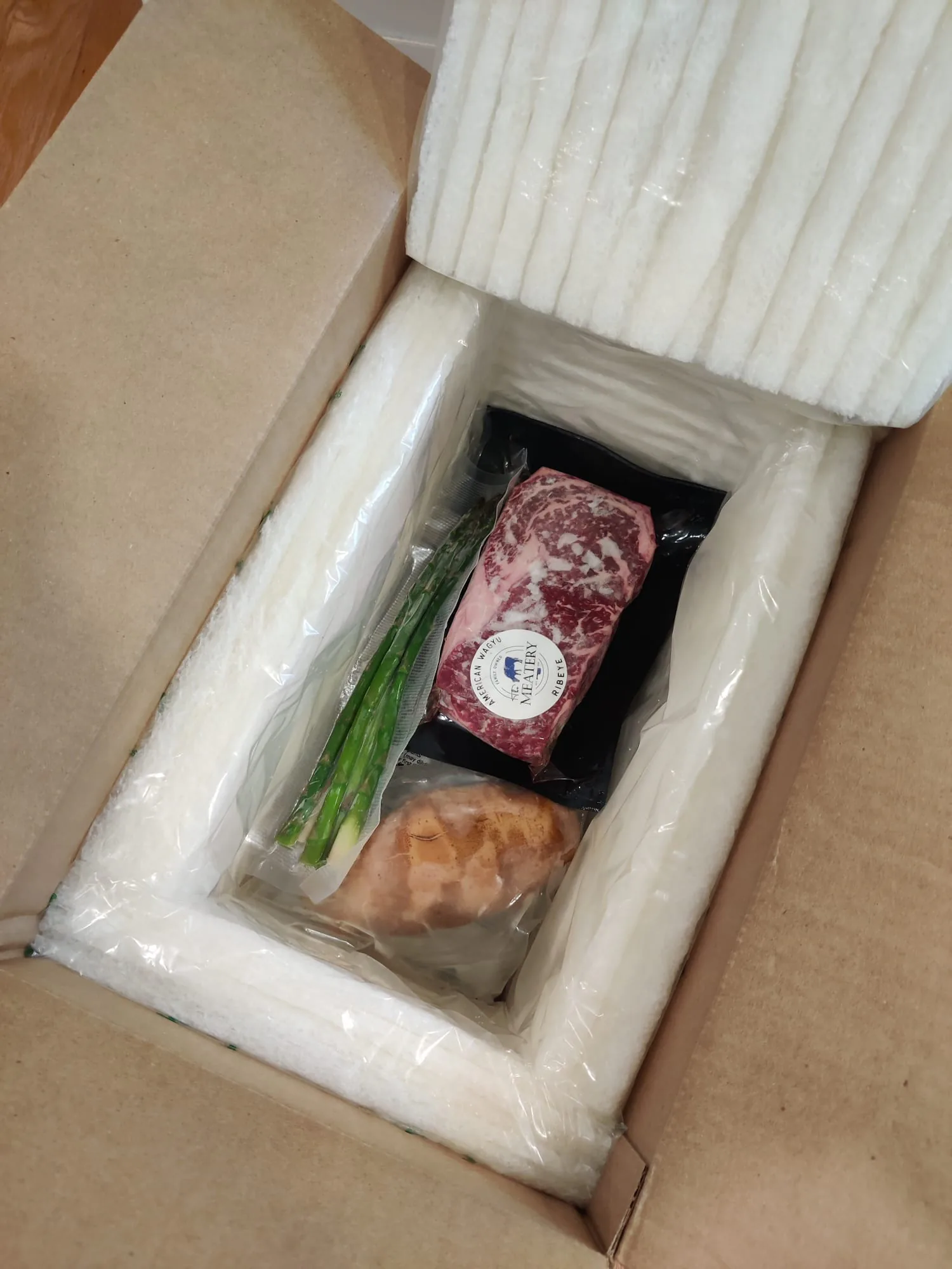 Open shipping box with an insulated liner holding a vacuum-sealed wagyu steak, scallions and produce