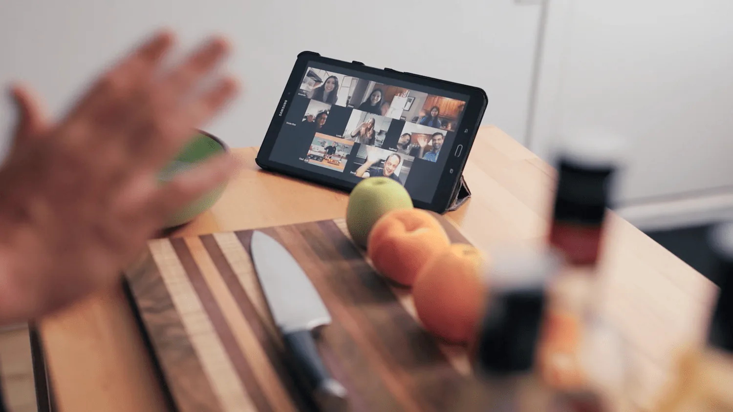 Hand waving at a tablet propped on a kitchen counter showing a grid of video call participants