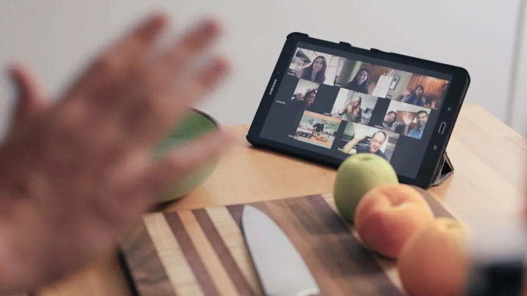 Hand waving at a tablet on a kitchen counter that shows a grid of people on a video call