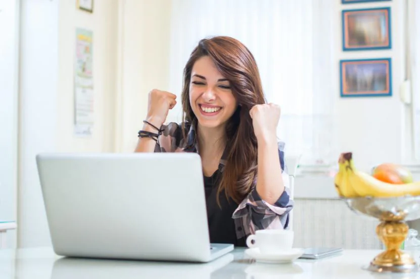 Smiling woman raising both fists in celebration in front of an open laptop at home