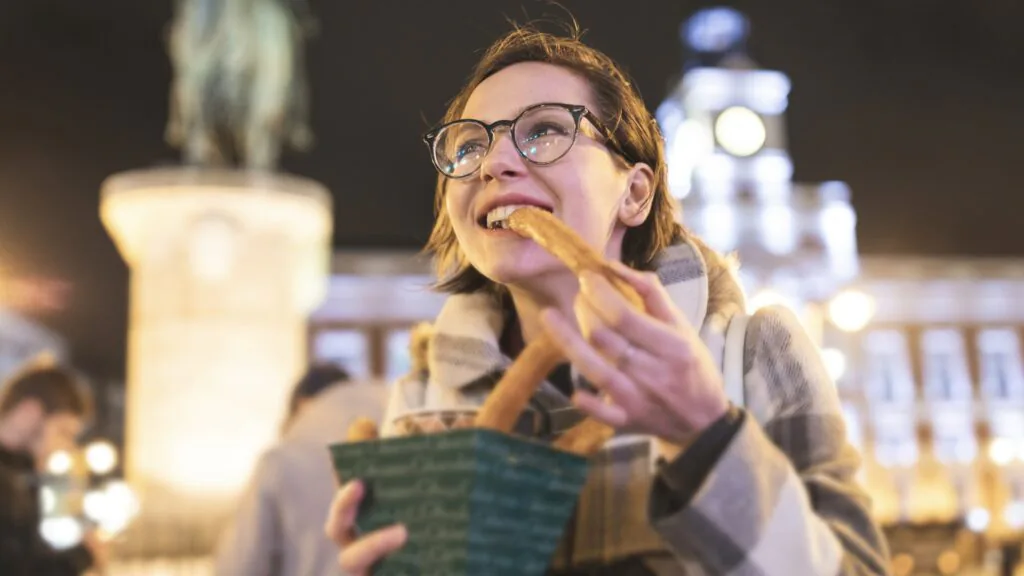 Smiling woman eating a churro from a paper cone in a lit city square at night