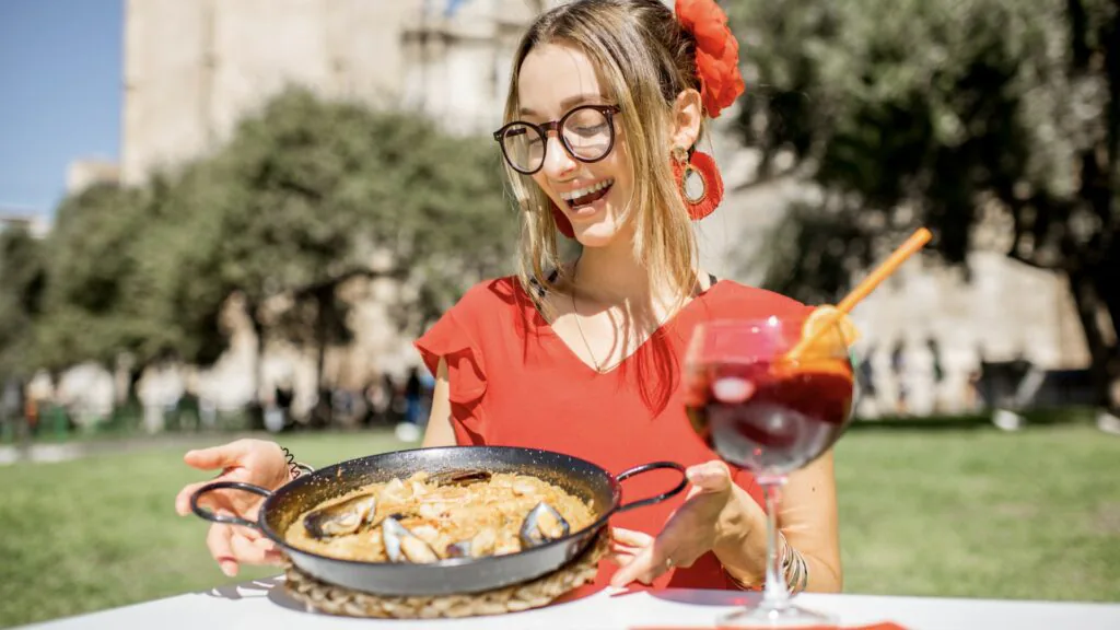 Woman in a red dress holding a pan of seafood paella and a glass of sangria outdoors