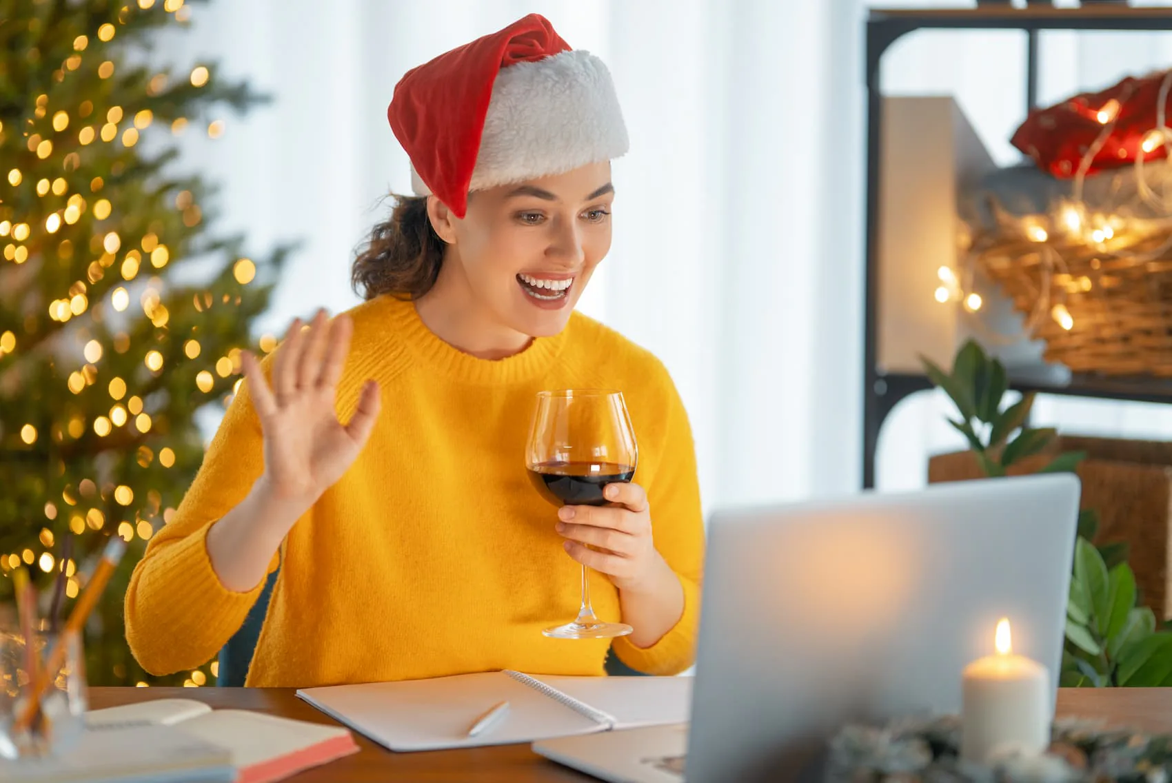 Woman in a Santa hat waving at her laptop with a glass of red wine by a lit Christmas tree