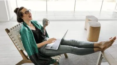 Woman laughing with a laptop on her lap and a mug in hand while lounging in a chair at home