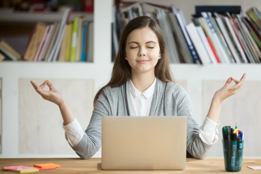 Woman sitting at her desk behind a laptop with eyes closed and hands in a meditation pose