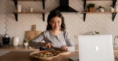 Woman smiling as she slices food on a wooden board at her kitchen counter beside an open laptop