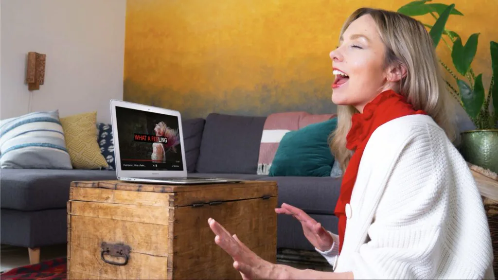 Woman singing along to a laptop on a wooden chest in her living room during a virtual karaoke session
