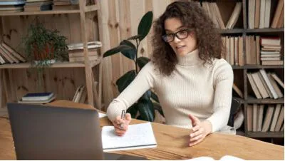 Woman in glasses taking notes next to a laptop at a wooden desk in a book-lined room