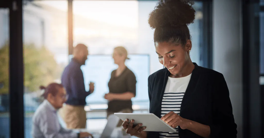 Woman smiling as she works on a tablet in an open-plan office with colleagues behind her
