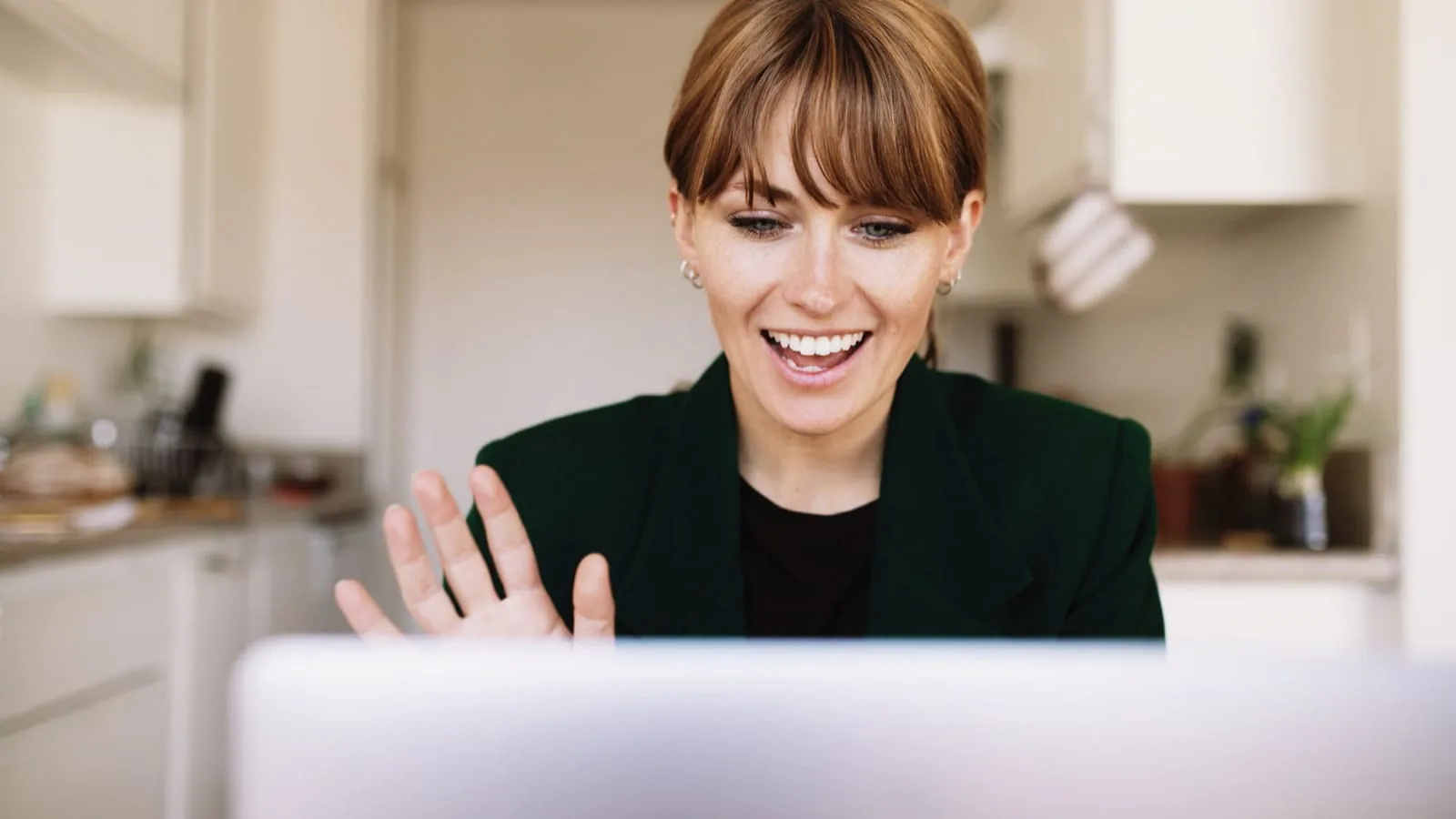 Smiling woman waving at her laptop screen during a video call in her kitchen