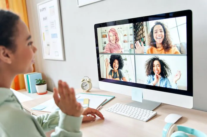 Woman waving at a desktop monitor showing four colleagues smiling on a video call