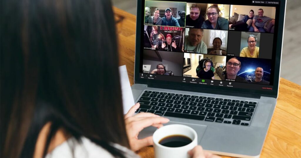 Woman holding a coffee mug watches a laptop showing a video call grid of a dozen smiling colleagues