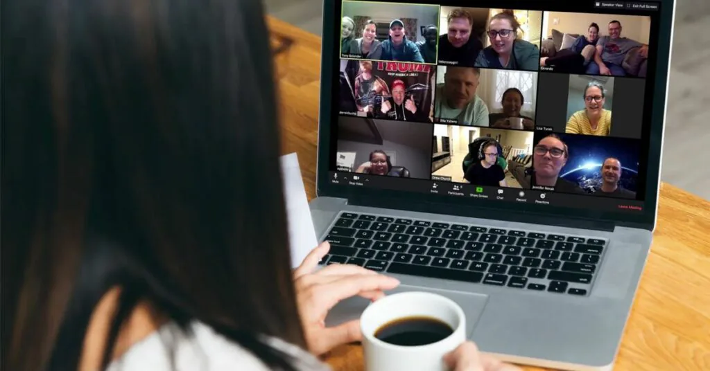 Woman holding a coffee mug watches a laptop showing a video call grid of a dozen smiling colleagues