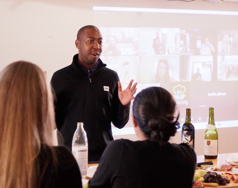 Host speaking to a seated team while remote colleagues appear on a projected video-call grid