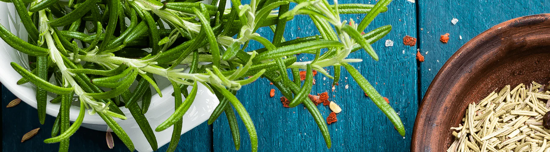 Fresh rosemary sprigs in a white bowl on a blue wooden table with peppercorns and a bowl of seeds