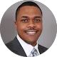 Byron Hawkins smiling in a dark suit and patterned tie against a grey studio backdrop