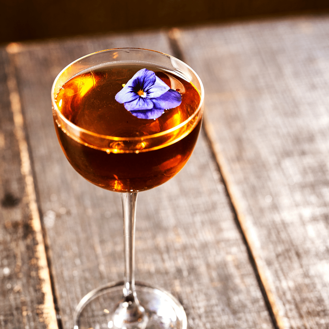 Amber cocktail in a coupe glass garnished with a blue edible flower on a bar top