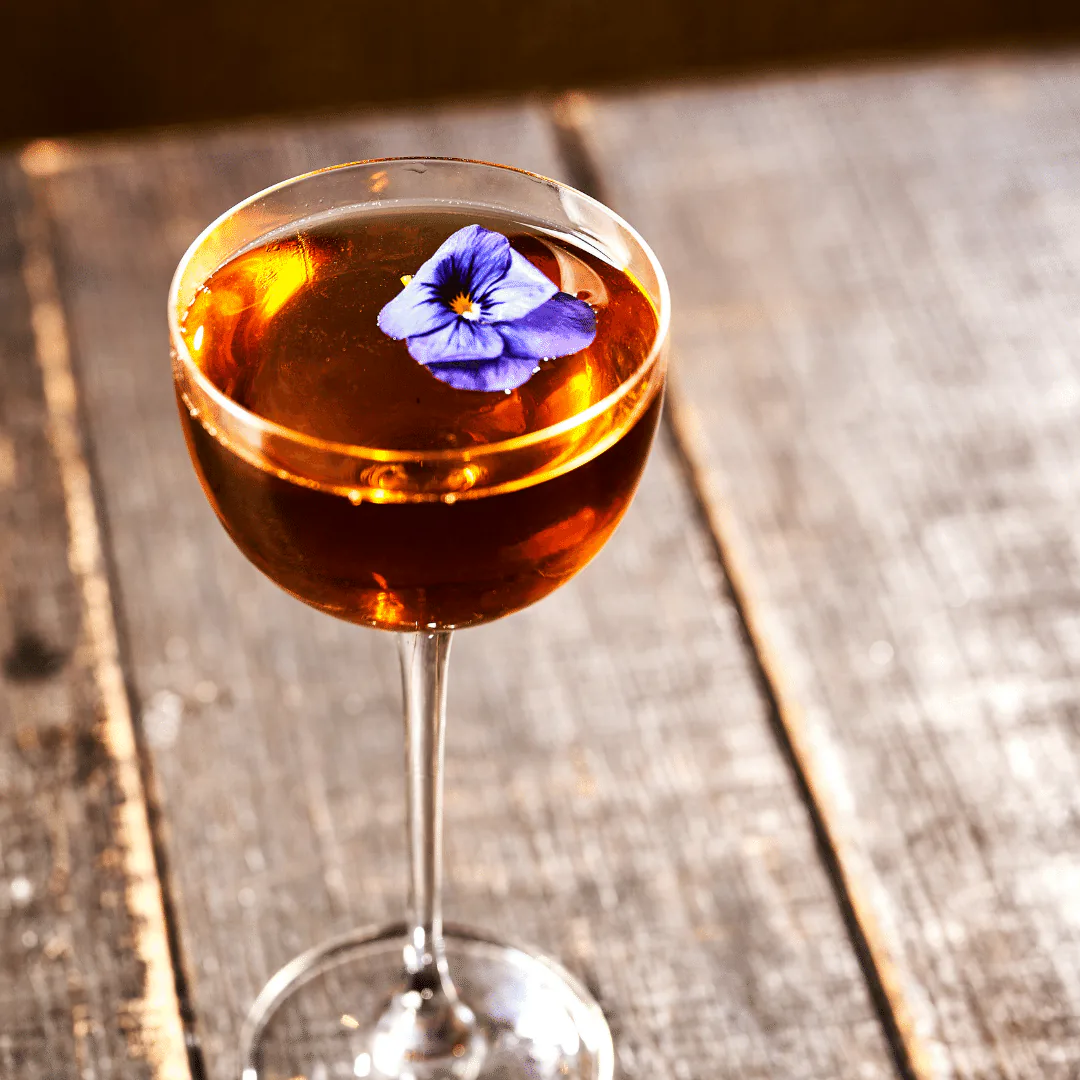Amber cocktail in a coupe glass garnished with a blue edible flower on a bar top
