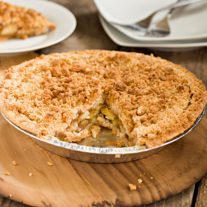 Apple pie with a golden crumble topping in a tin with one slice cut out, on a wooden board