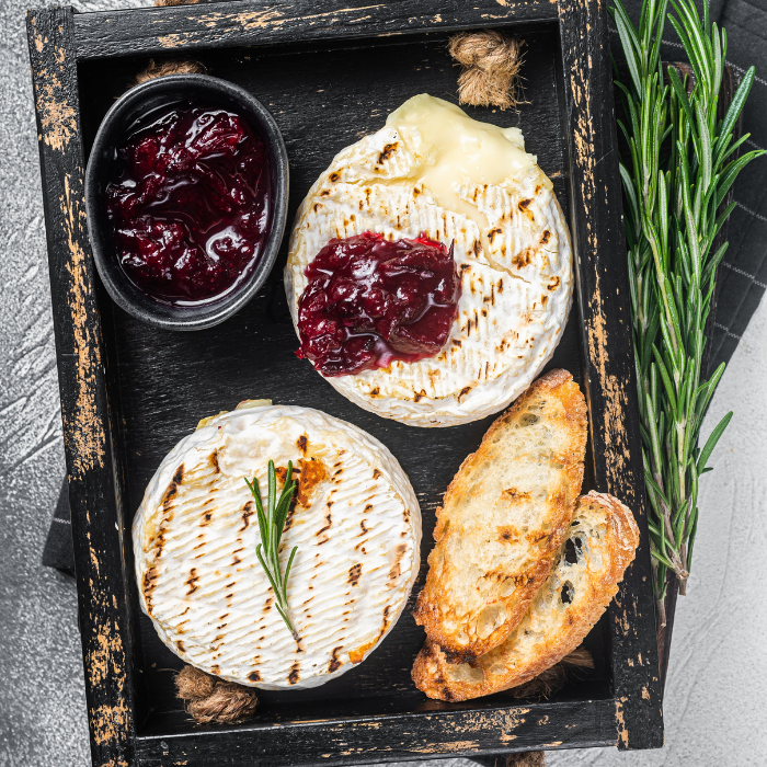 Baked soft cheese rounds topped with cranberry sauce and rosemary in a dark tray with toasted bread