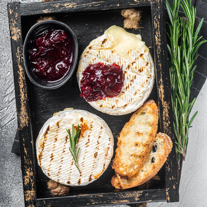 Baked soft cheese rounds topped with cranberry sauce and rosemary in a dark tray with toasted bread