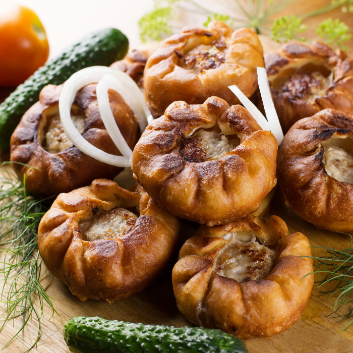 Pile of golden baked pastries filled with meat, with dill, onion rings and cucumbers alongside