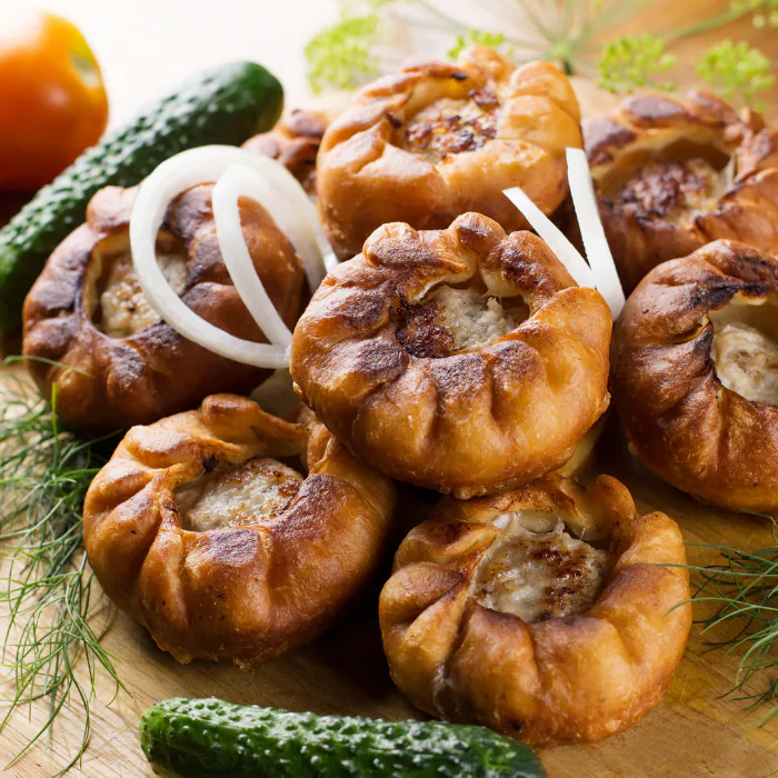 Pile of golden baked pastries filled with meat, with dill, onion rings and cucumbers alongside