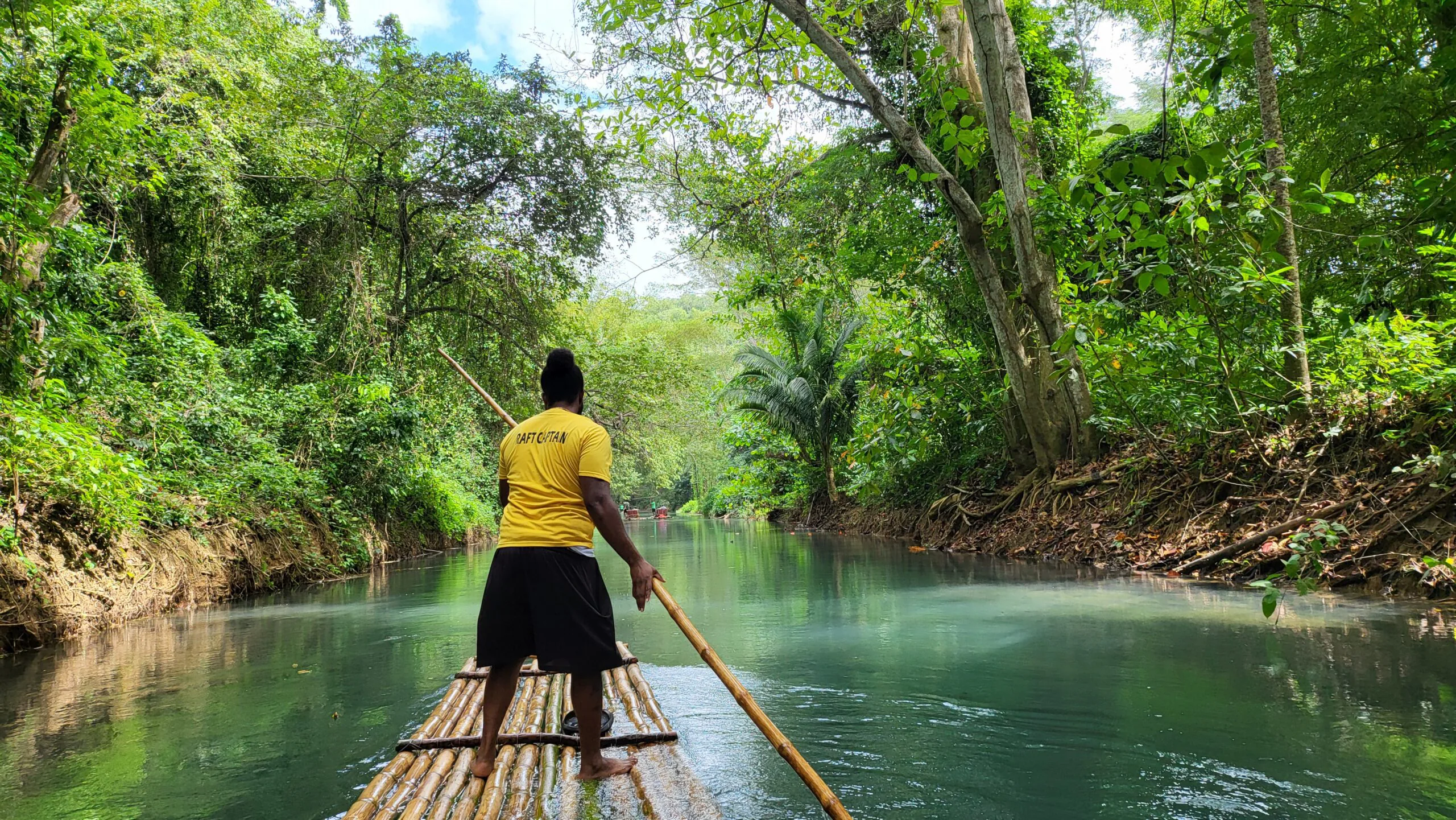 Guide poling a bamboo raft down a calm jungle river lined with tropical greenery