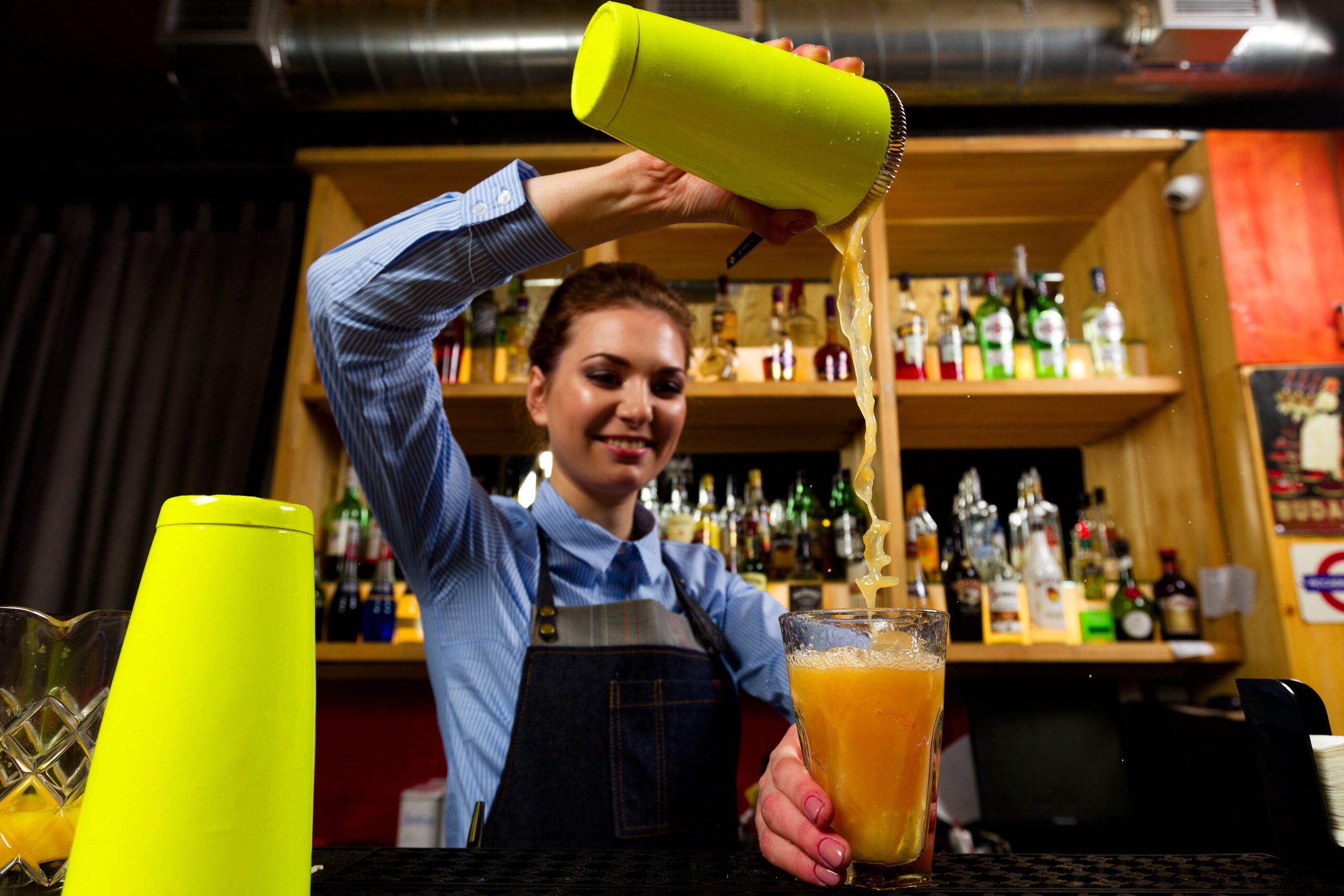 Bartender pouring a cocktail from a green shaker into a tall glass in front of a stocked back bar