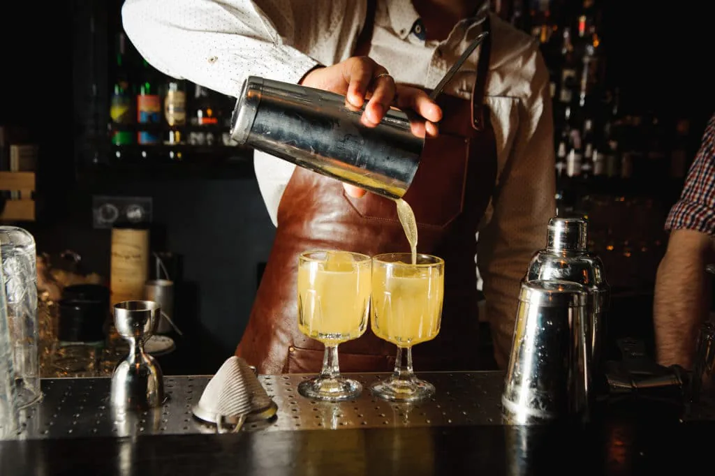 Bartender in a leather apron pouring a yellow cocktail from a shaker into two coupe glasses