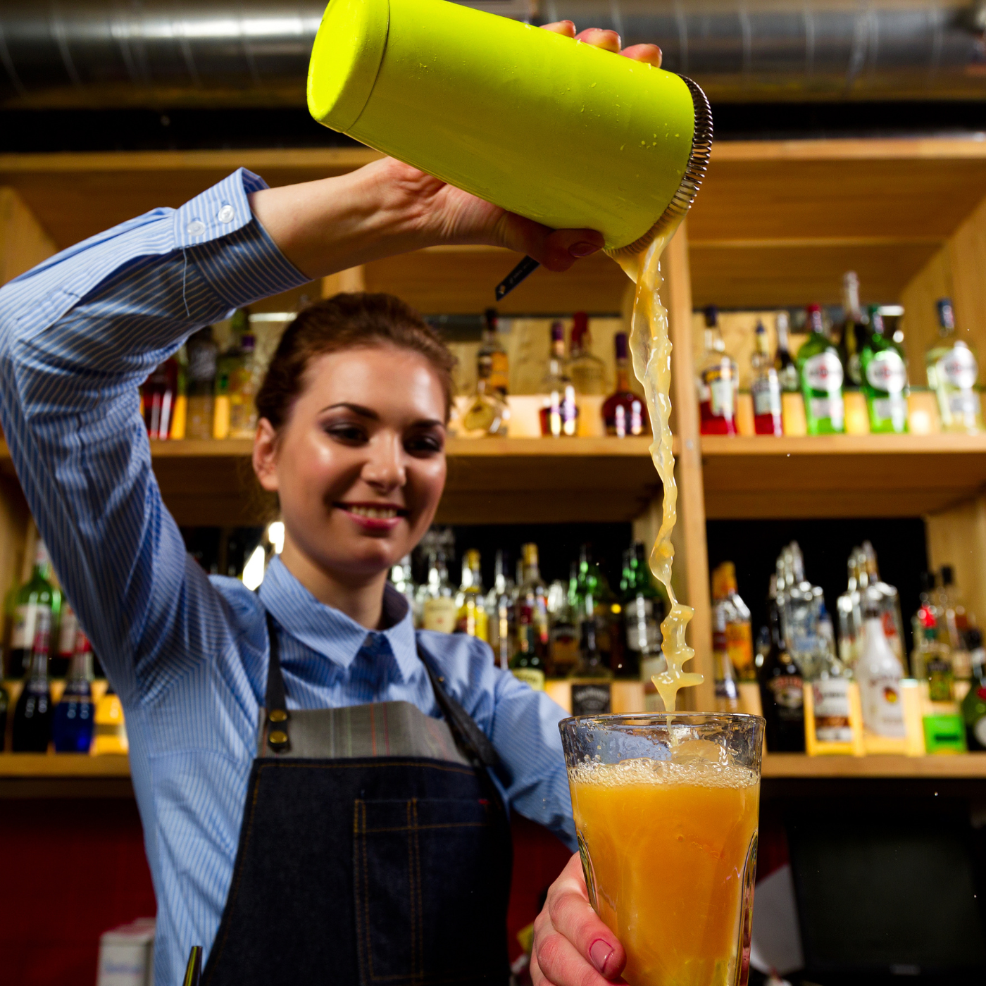 Bartender pouring a bright orange cocktail from a yellow shaker into a glass in front of a lit back bar