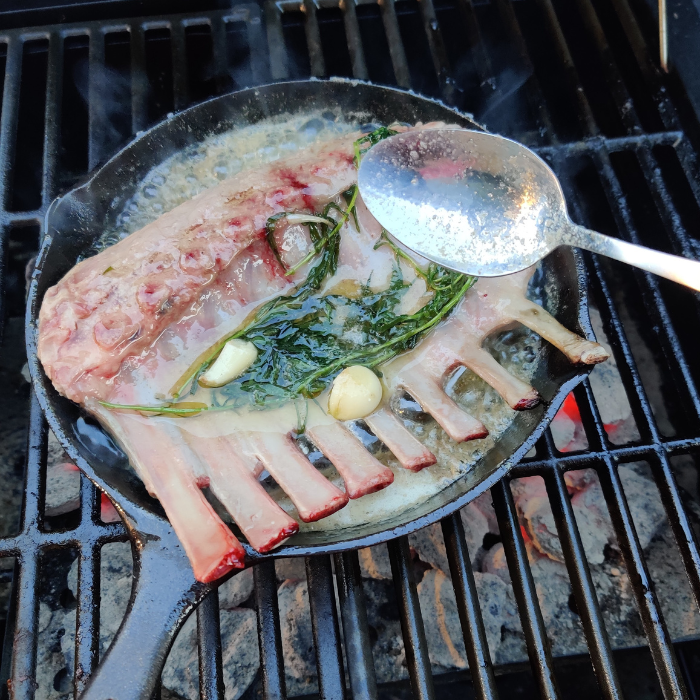Rack of lamb basting in herb butter in a cast iron skillet on a grill