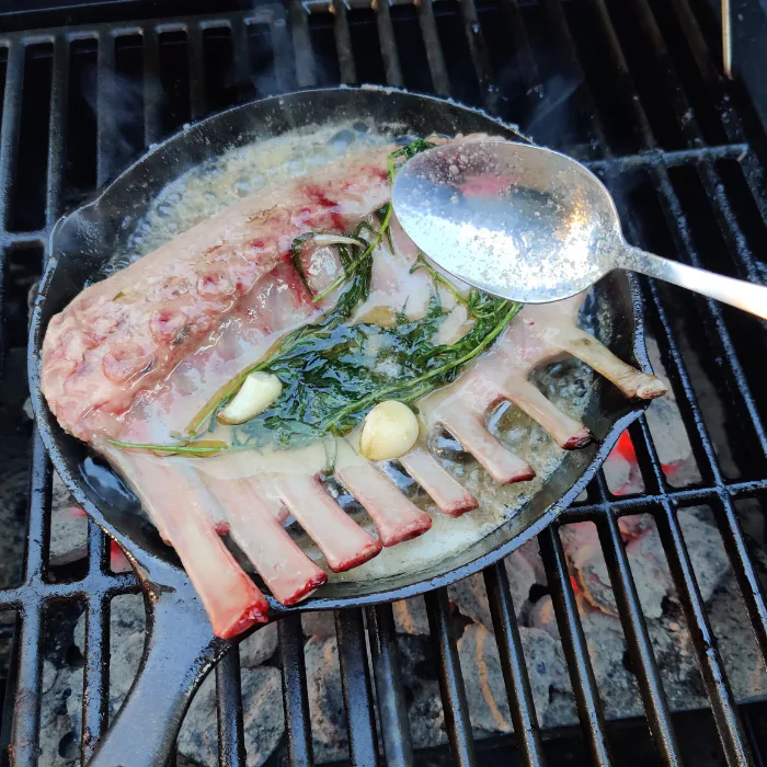 Rack of lamb basting in herb butter in a cast iron skillet on a grill