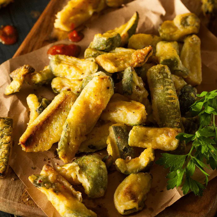 Golden battered zucchini fries piled on parchment paper with parsley and cherry tomatoes