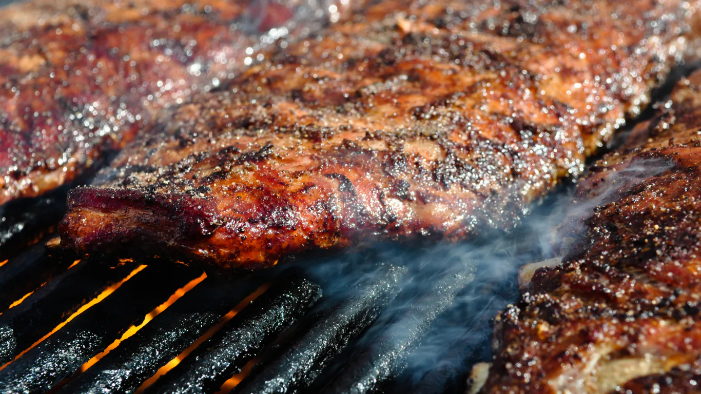 Racks of barbecue ribs searing over glowing coals with smoke rising from the grill