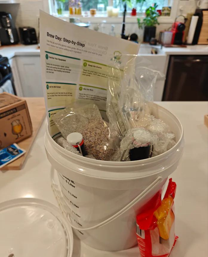 Beer brewing kit packed into a white bucket with grain, hops and tubing and a Brew Day step-by-step sheet