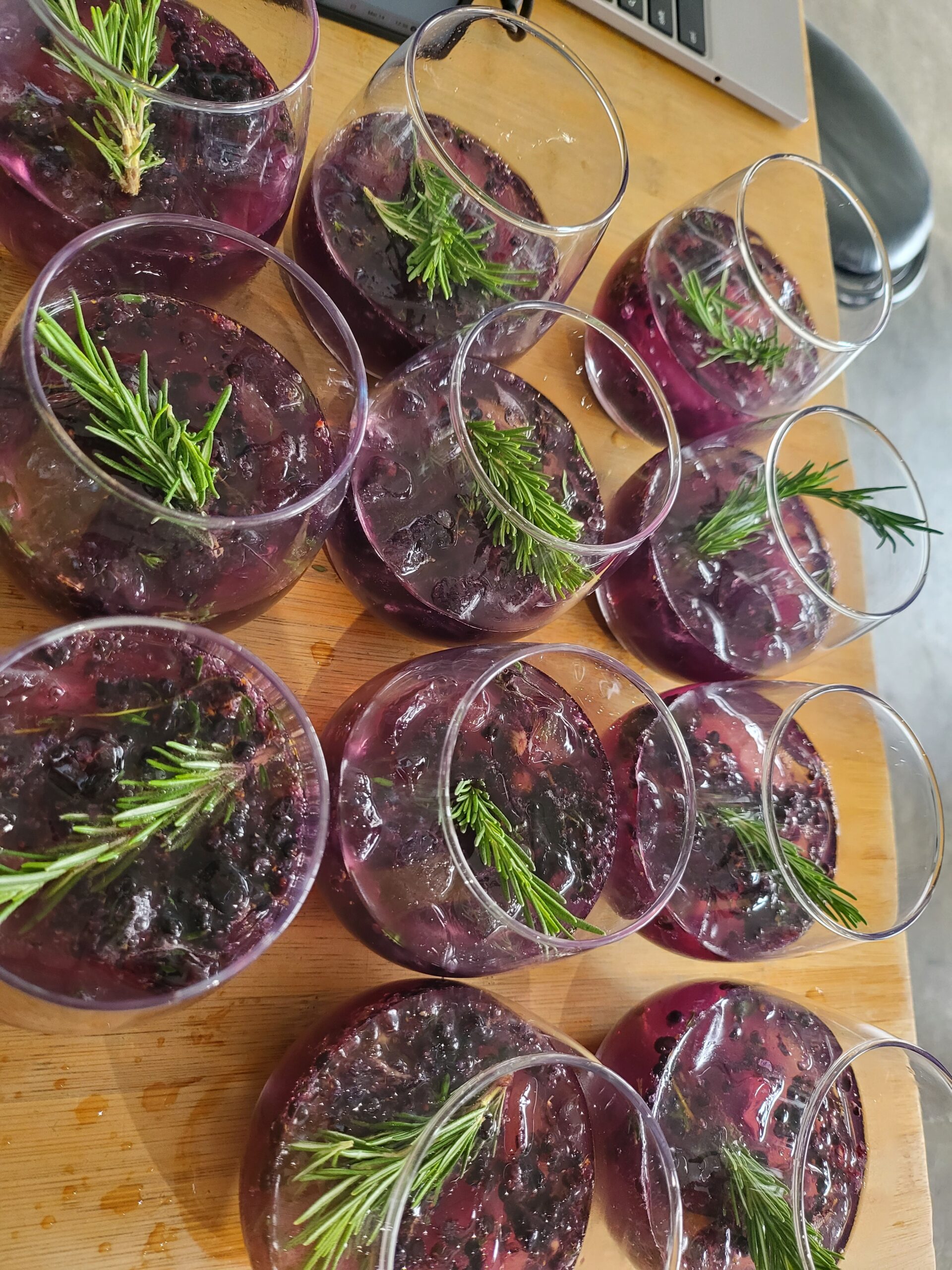 Rows of glasses filled with a dark berry cocktail, each garnished with a fresh rosemary sprig