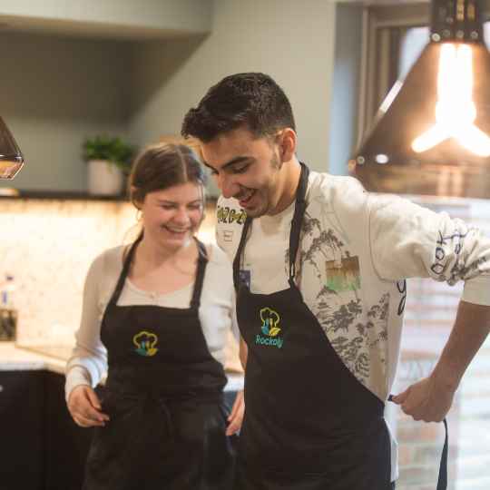 Two people in black aprons embroidered with the Rockoly chef logo, smiling in a kitchen