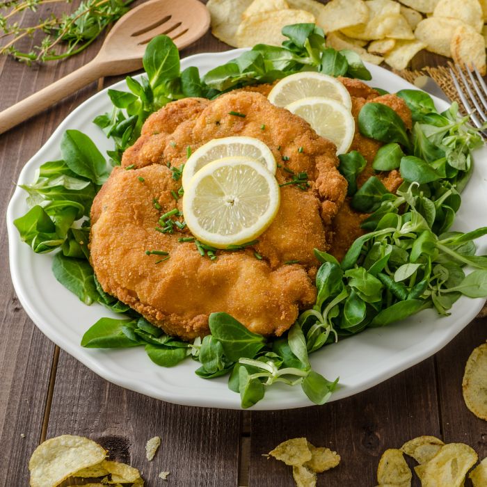 Golden breaded schnitzel topped with lemon slices on a bed of green leaves with potato chips
