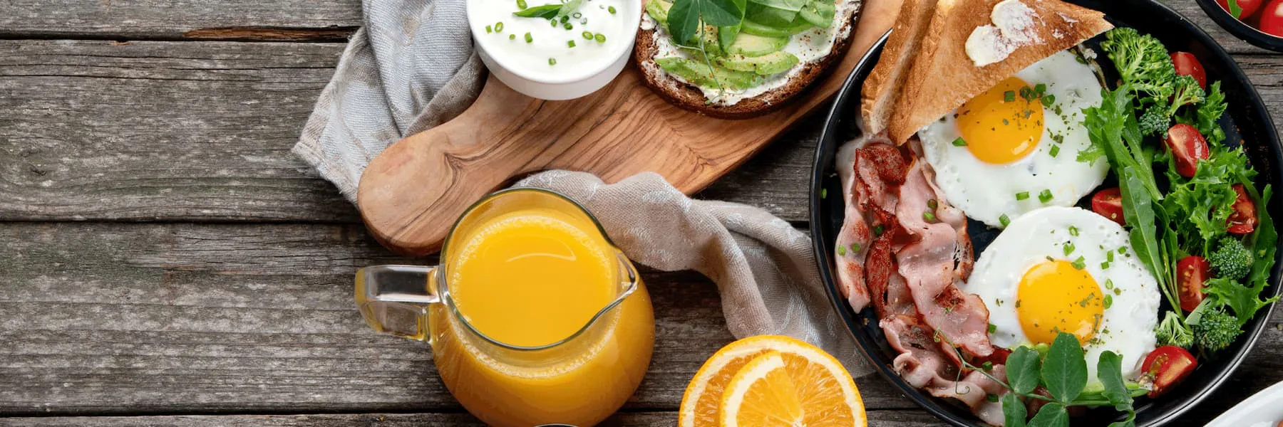 Brunch spread of fried eggs and bacon in a skillet, avocado toast, salad and a jug of orange juice
