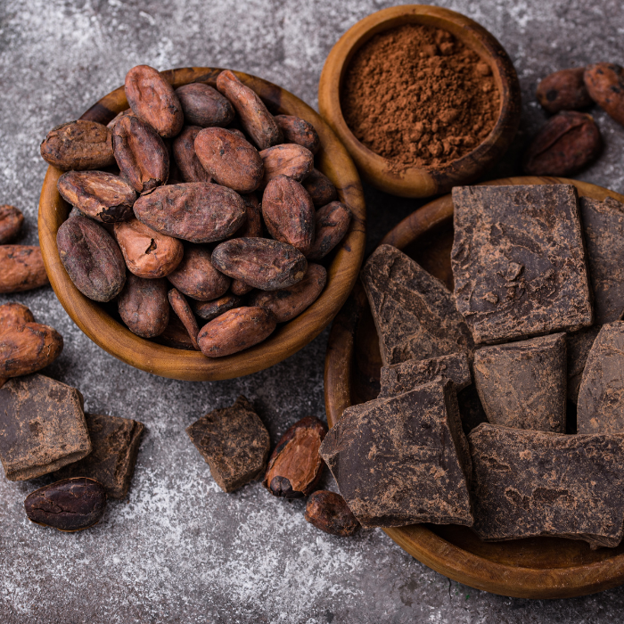 Cacao beans and cocoa powder in wooden bowls beside chunks of raw dark chocolate