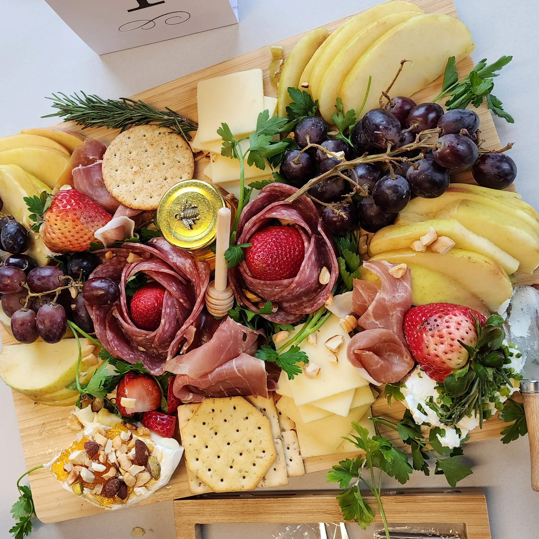 Grazing board of salami roses, prosciutto, sliced cheeses, strawberries, grapes, crackers and a honey jar