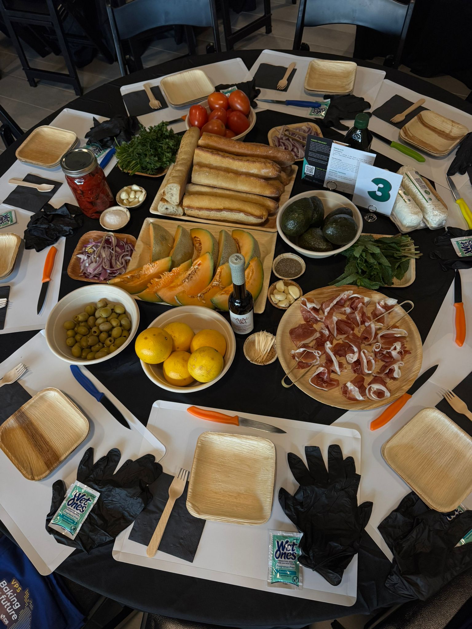 Round table spread with bread, prosciutto, olives, melon, lemons and herbs beside plates and gloves