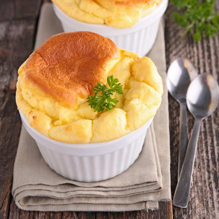 Golden puffed cheddar soufflés in white ramekins garnished with parsley, with spoons on a linen napkin