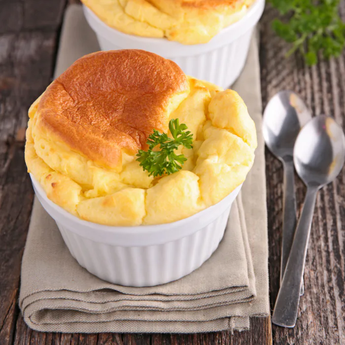 Golden puffed cheddar soufflés in white ramekins garnished with parsley, with spoons on a linen napkin